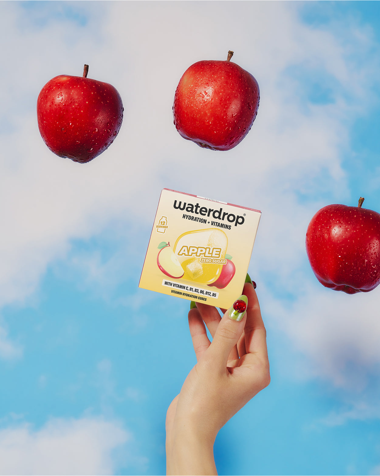 Mano sosteniendo una pequeña caja con ilustración de manzana, cielo azul y tres manzanas rojas flotando.