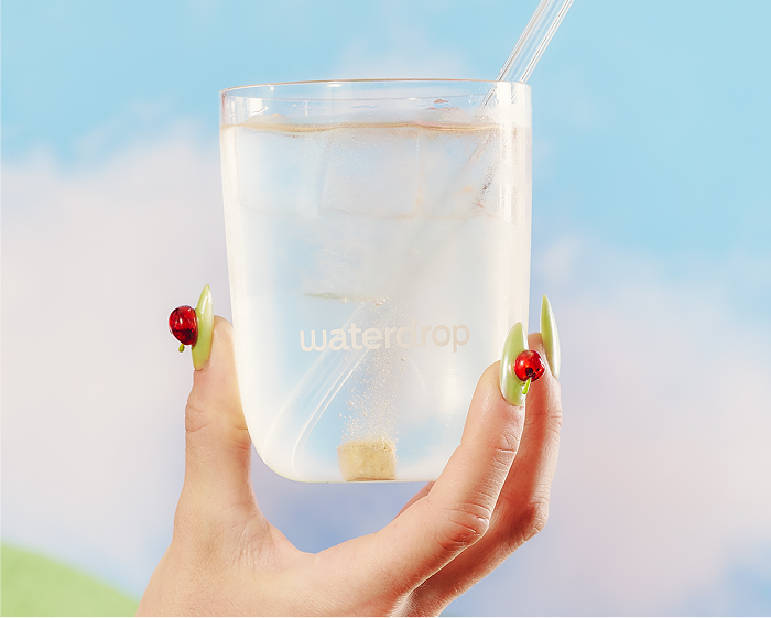 Una mano sostiene un vaso transparente con agua y hielo, con una pajilla, contra un cielo azul.